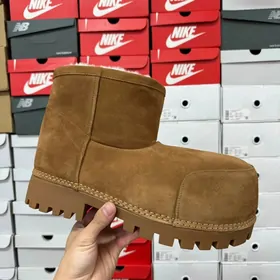 A chestnut suede mini boot with a lug sole held up in front of shoeboxes