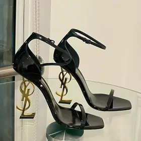 Black patent ankle-strap sandals with a sculpted gold heel on glass